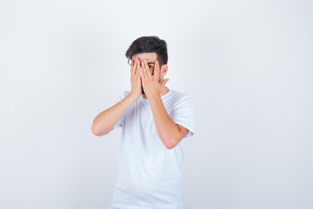 Jeune homme regardant à travers les doigts en t-shirt blanc et ayant l'air effrayé