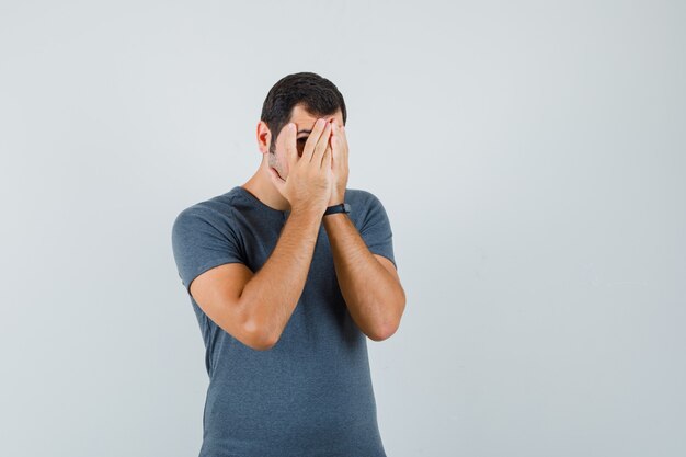 Jeune homme regardant à travers les doigts en t-shirt gris et à la peur. vue de face.