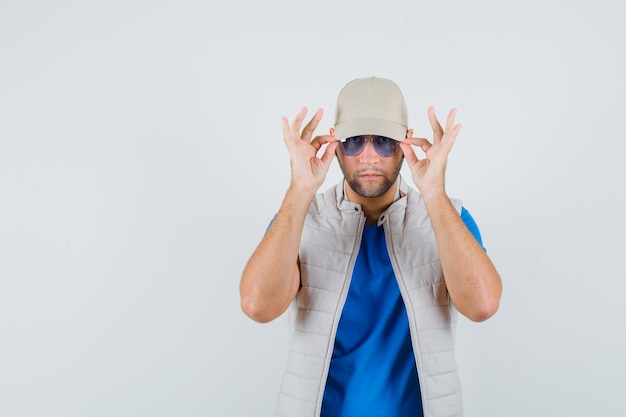 Jeune homme regardant à travers des lunettes en t-shirt, veste, casquette, vue de face.