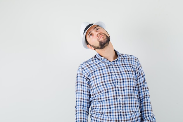 Jeune homme regardant vers le haut en chemise à carreaux, chapeau et à la recherche concentrée. vue de face.