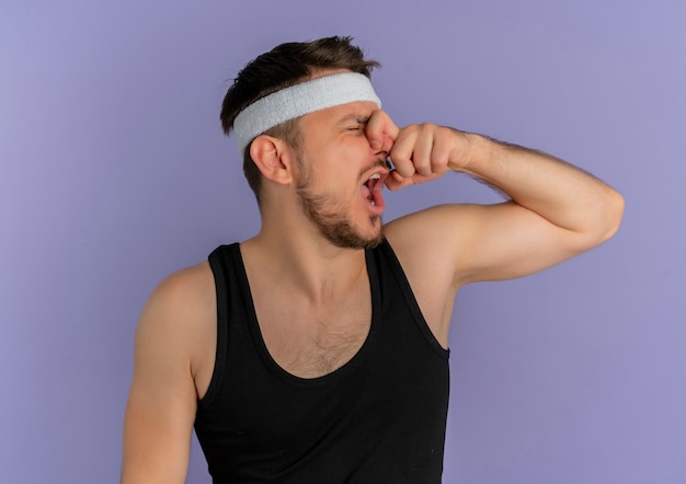 Jeune homme de remise en forme avec bandeau fermant son nez avec les doigts souffrant de puanteur debout sur un mur violet