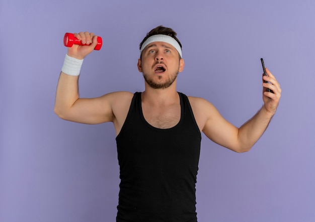 Jeune homme de remise en forme avec bandeau prenant selfie à l'aide de son smartphone posant avec haltère à la main debout sur le mur violet