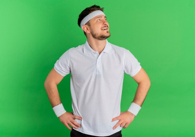 Jeune homme de remise en forme en chemise blanche avec bandeau à côté heureux et positif souriant confiant debout sur fond vert