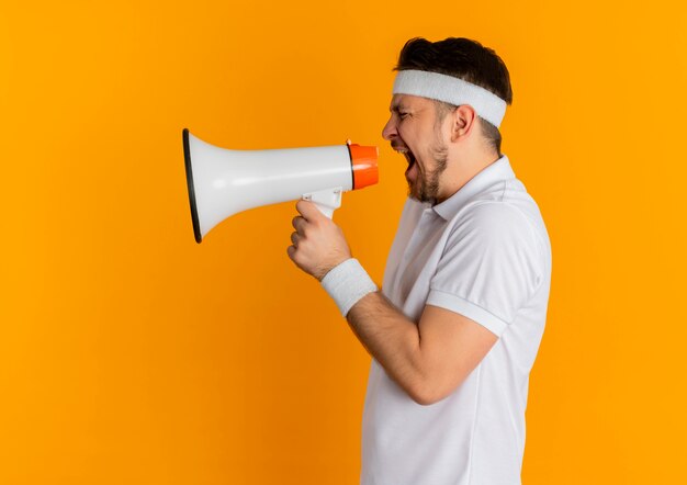 Jeune homme de remise en forme en chemise blanche avec bandeau criant au mégaphone avec une expression agressive debout sur un mur orange