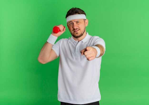 Jeune homme de remise en forme en chemise blanche avec bandeau faisant des exercices avec haltère pointant avec le doigt à la caméra debout sur fond vert