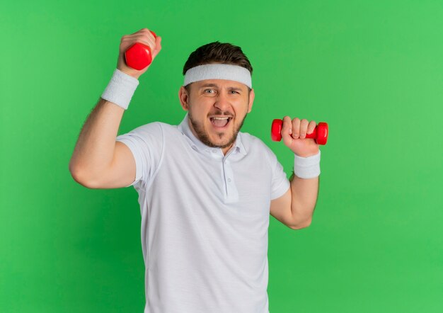 Jeune homme de remise en forme en chemise blanche avec bandeau faisant des exercices avec des haltères à la confusion debout sur fond vert