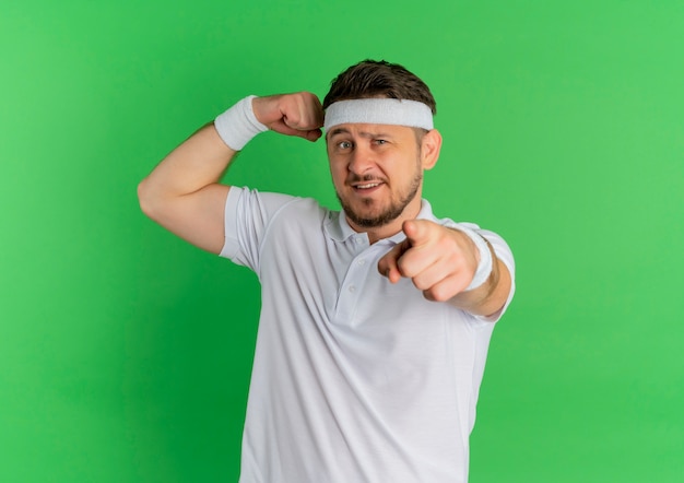 Jeune homme de remise en forme en chemise blanche avec bandeau levant le poing montrant les biceps, pointant du doigt vers l'avant, debout sur le mur vert