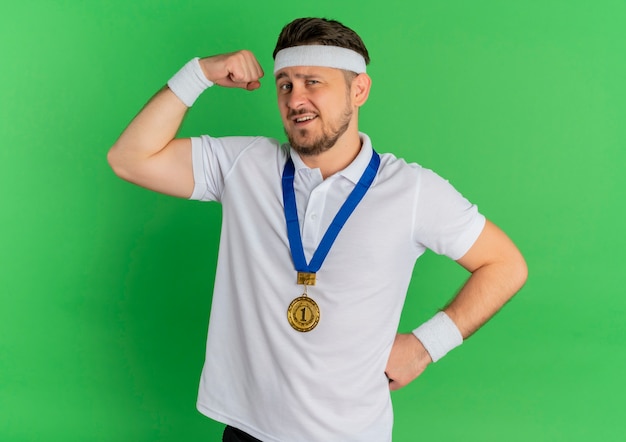 Jeune Homme De Remise En Forme En Chemise Blanche Avec Bandeau Et Médaille D'or Autour Du Cou Levant Le Poing Montrant Les Biceps, Concept Gagnant Debout Sur Fond Vert