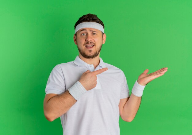 Jeune homme de remise en forme en chemise blanche avec bandeau pointant avec le doigt sur le côté présentant le bras de sa main debout sur le mur vert