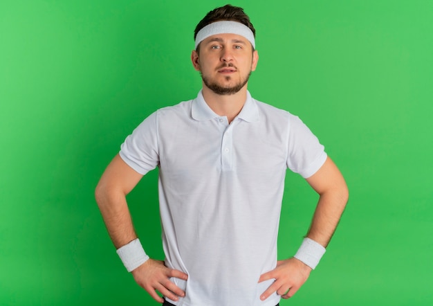 Jeune homme de remise en forme en chemise blanche avec bandeau regardant la caméra avec une expression confiante debout sur fond vert