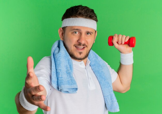 Jeune homme de remise en forme en chemise blanche avec bandeau et serviette autour du cou travaillant avec haltère regardant la caméra avec un sourire amical debout sur fond vert