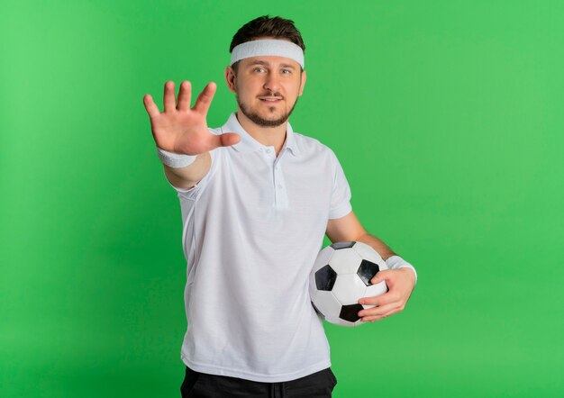 Jeune homme de remise en forme en chemise blanche avec bandeau tenant un ballon de football regardant la caméra avec les bras levés debout sur fond vert