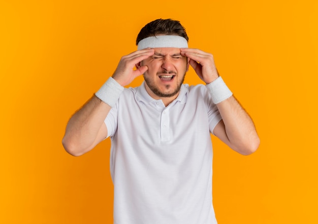 Jeune homme de remise en forme en chemise blanche avec bandeau touchant les tempes à la souffrance souffrant de douleur debout sur le mur orange