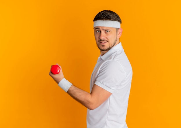 Jeune homme de remise en forme en chemise blanche avec bandeau travaillant avec haltère à la confiance debout sur le mur orange