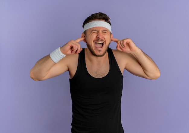 Jeune homme de remise en forme avec serre-tête fermant les oreilles avec les doigts avec une expression agacée debout sur un mur violet