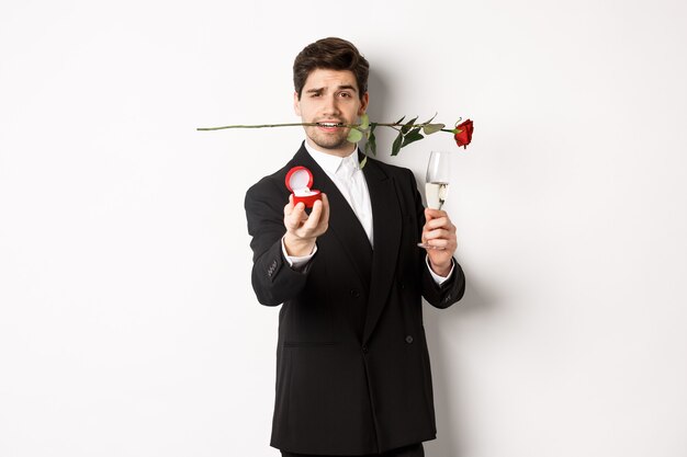 Jeune homme romantique en costume faisant une proposition, tenant une rose dans les dents et une coupe de champagne, montrant une bague de fiançailles, demandant à l'épouser, debout sur fond blanc.