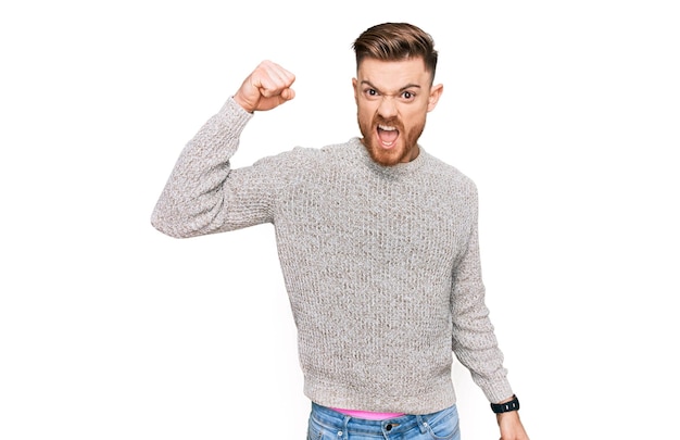 Jeune homme rousse portant un pull d'hiver décontracté en colère et fou levant le poing frustré et furieux en criant de colère. rage et concept agressif.