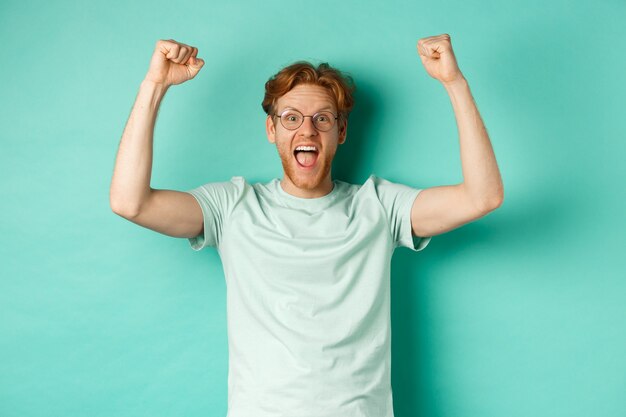 Jeune homme rousse se sentant comme un champion, levant les mains dans le geste de la pompe de poing et criant oui de joie, gagnant le prix, triomphant du succès, debout sur fond de menthe.