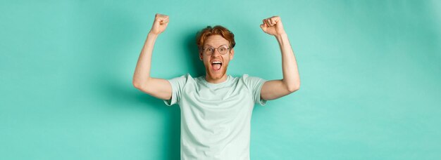 Jeune homme rousse se sentant comme un champion levant les mains en geste de pompe de poing et criant oui avec