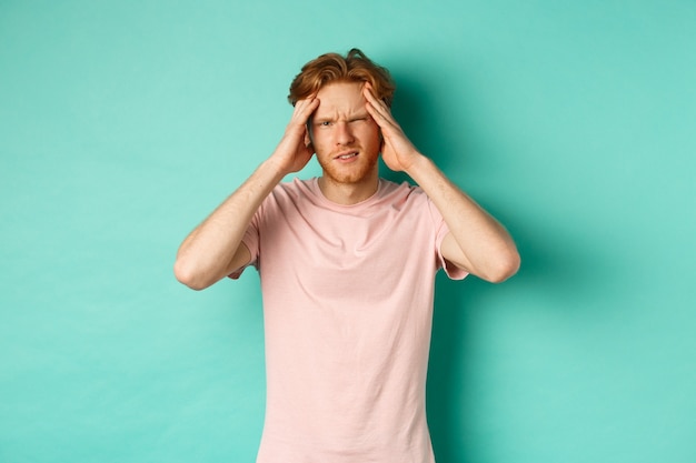 Jeune homme roux touchant la tête et ayant l'air étourdi, ressentant des maux de tête ou des migraines, debout en t-shirt sur fond menthe.