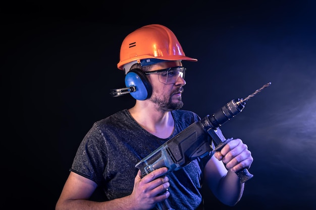 Un jeune homme en salopette casque de protection casque tenir une perceuse électrique