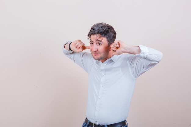 Jeune homme se branchant les oreilles avec les doigts en chemise blanche, jeans et à la peur