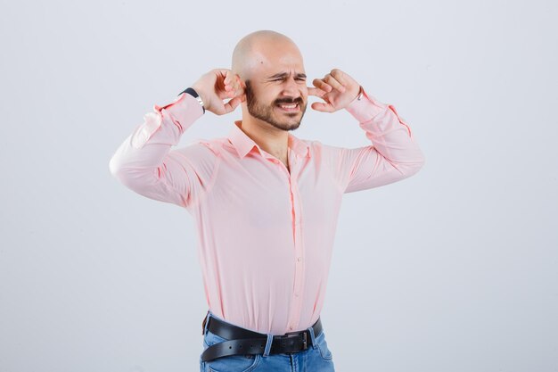 Jeune homme se branchant les oreilles avec les doigts tout en devenant fou en chemise rose, jeans, vue de face.