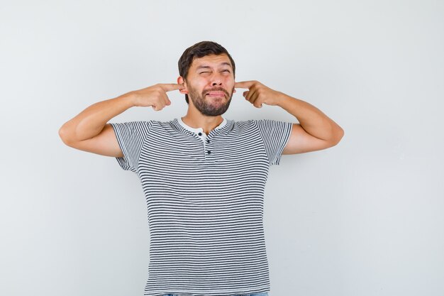 Jeune homme se branchant les oreilles avec les doigts tout en faisant la grimace en t-shirt rayé et l'air ennuyé, vue de face.