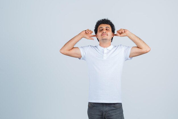 Jeune homme se branchant les oreilles avec l'index, fermant les yeux en t-shirt blanc et en jean et ayant l'air sérieux, vue de face.