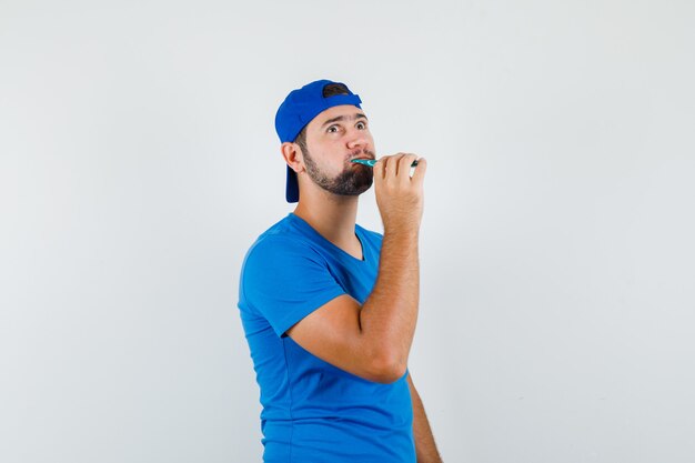 Jeune homme se brosser les dents tout en regardant en t-shirt bleu et casquette et à la pensif