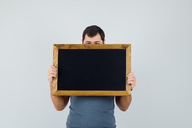 Jeune homme se cachant le visage derrière le tableau noir en t-shirt gris