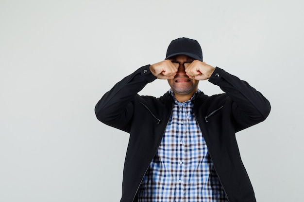 Jeune homme se frottant les yeux en pleurant en chemise, veste, casquette et à l'offensé, vue de face.