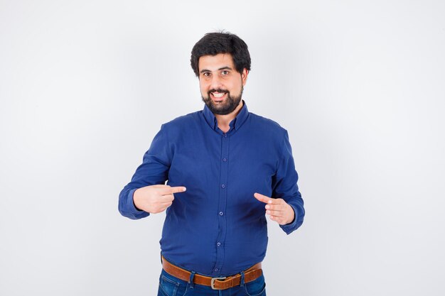 Jeune homme se pointant en chemise bleue et jeans et à l'optimisme. vue de face.