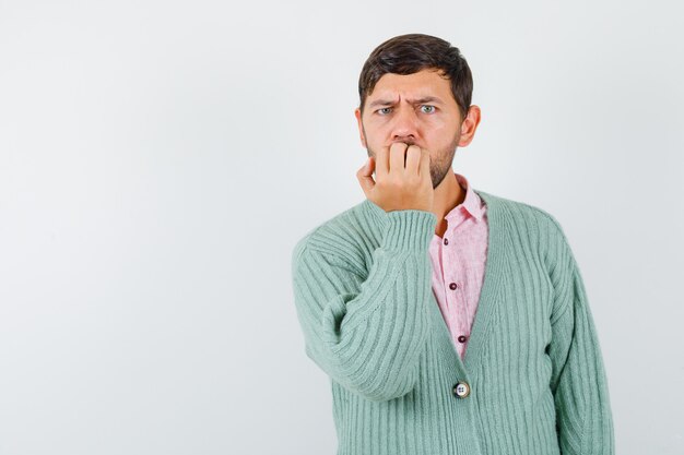 Jeune homme se rongeant les ongles avec émotion en chemise, cardigan et semblant anxieux. vue de face.