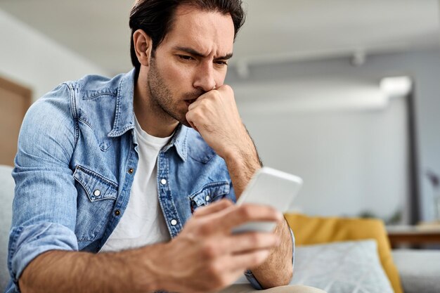 Jeune homme se sentant inquiet en lisant un message texte sur un téléphone portable à la maison