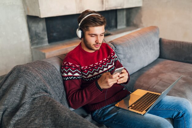 Jeune homme séduisant sur le canapé à la maison en hiver avec smartphone dans les écouteurs, écouter de la musique, porter un pull en tricot rouge, travaillant sur ordinateur portable, pigiste