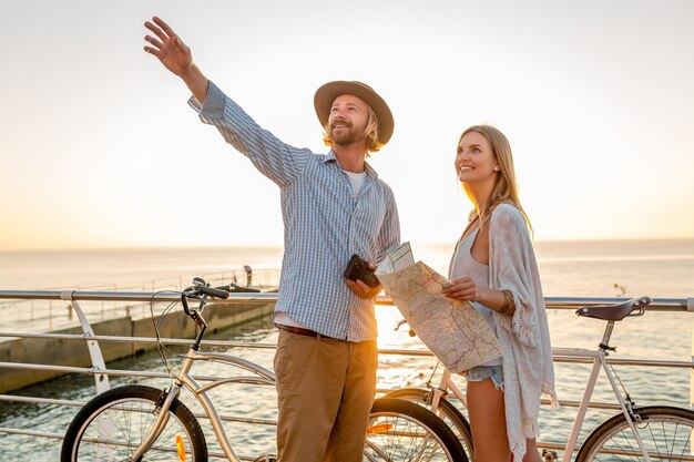 Jeune homme séduisant et femme voyageant à bicyclette, tenant la carte et le tourisme, couple romantique en vacances d'été au bord de la mer au coucher du soleil, amis s'amusant ensemble