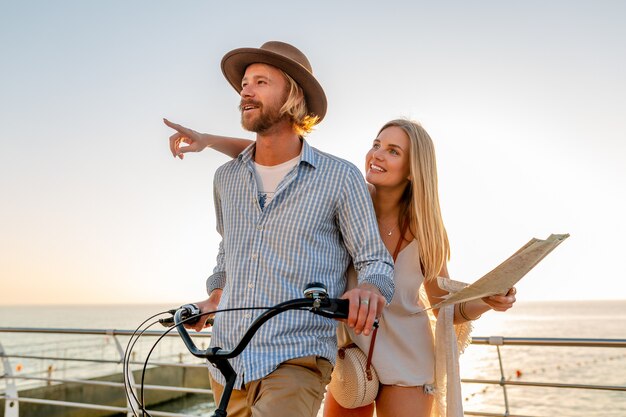 Jeune homme séduisant et femme voyageant à bicyclette, tenant la carte, visites touristiques, couple romantique en vacances d'été au bord de la mer au coucher du soleil, tenue de style boho hipster, amis s'amusant ensemble