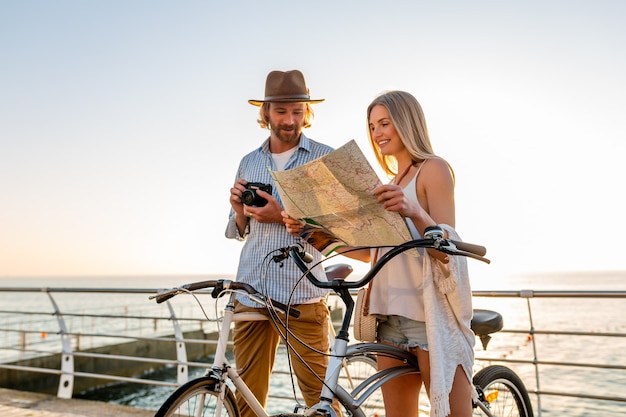 Jeune homme séduisant et femme voyageant sur des bicyclettes tenant une carte, tenue de style hipster, amis s'amusant ensemble, visites touristiques prenant une photo à la caméra, couple en vacances d'été sur la mer au coucher du soleil