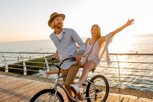 Jeune homme séduisant et femme voyageant à vélo, couple romantique en vacances d'été au bord de la mer au coucher du soleil, tenue de style boho hipster, amis s'amusant ensemble