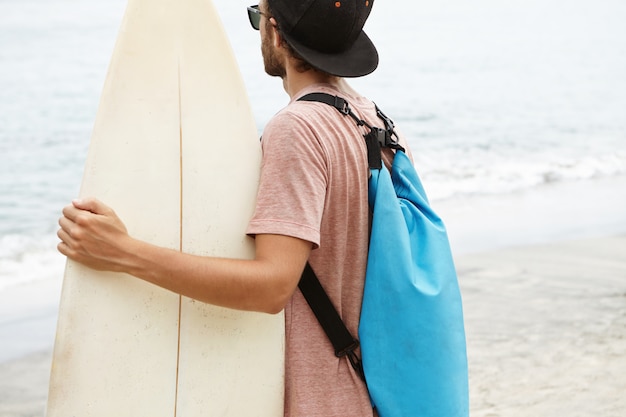 Jeune homme séduisant habillé avec désinvolture, portant snapback et lunettes de soleil, tenant sa planche de surf blanche et regardant la mer. Surfeur débutant se préparant à l'entraînement