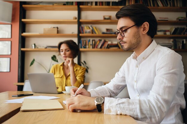 Jeune homme séduisant à lunettes assis à la table écrivant pensivement dans le bloc-notes avec un collègue en arrière-plan au travail dans un bureau moderne