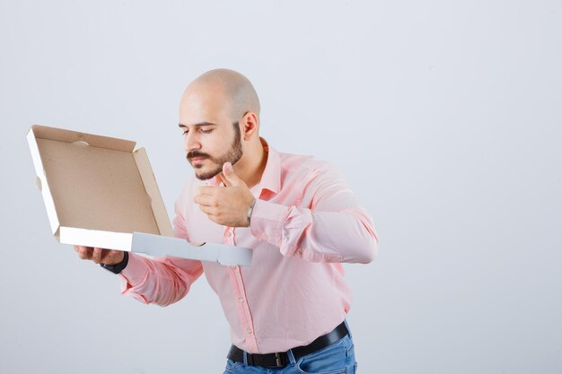 Jeune homme sentant une boîte à pizza ouverte en chemise, jeans et charmant, vue de face.