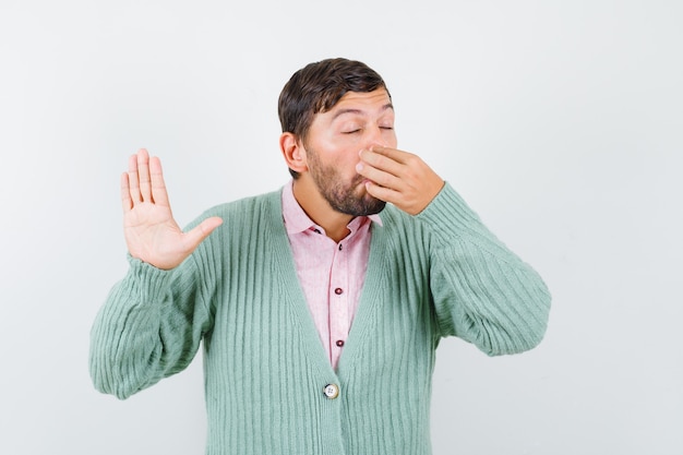 Photo gratuite jeune homme sentant quelque chose d'affreux, pinçant le nez, montrant un panneau d'arrêt en chemise, cardigan et ayant l'air dégoûté. vue de face.