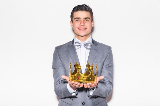 Photo gratuite jeune homme sérieux en costume-cravate, tenant une couronne d'or sur sa tête, sur un mur blanc