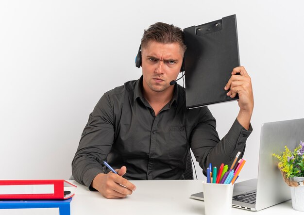 Jeune homme sérieux travailleur de bureau blonde sur les écouteurs est assis au bureau avec des outils de bureau à l'aide d'un ordinateur portable détient un stylo et un presse-papiers isolé sur fond blanc avec espace de copie