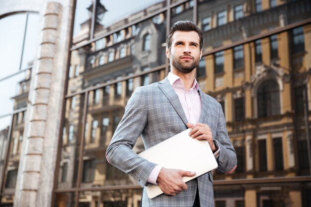 Jeune homme sérieux en veste tenant un ordinateur portable en position debout