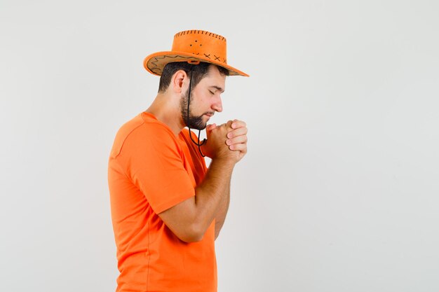 Jeune homme serrant les mains dans un geste de prière en t-shirt orange, chapeau et à l'air plein d'espoir.