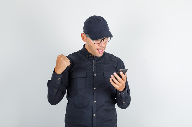 Jeune homme serrant le poing avec smartphone à la main en chemise noire et casquette