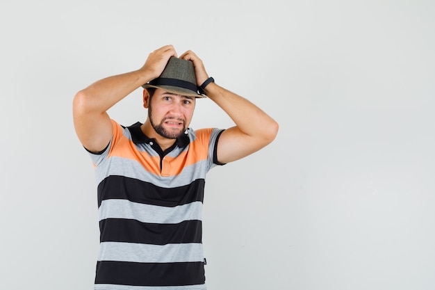 Photo gratuite jeune homme serrant la tête dans les mains en t-shirt, chapeau et regardant anxieux, vue de face.